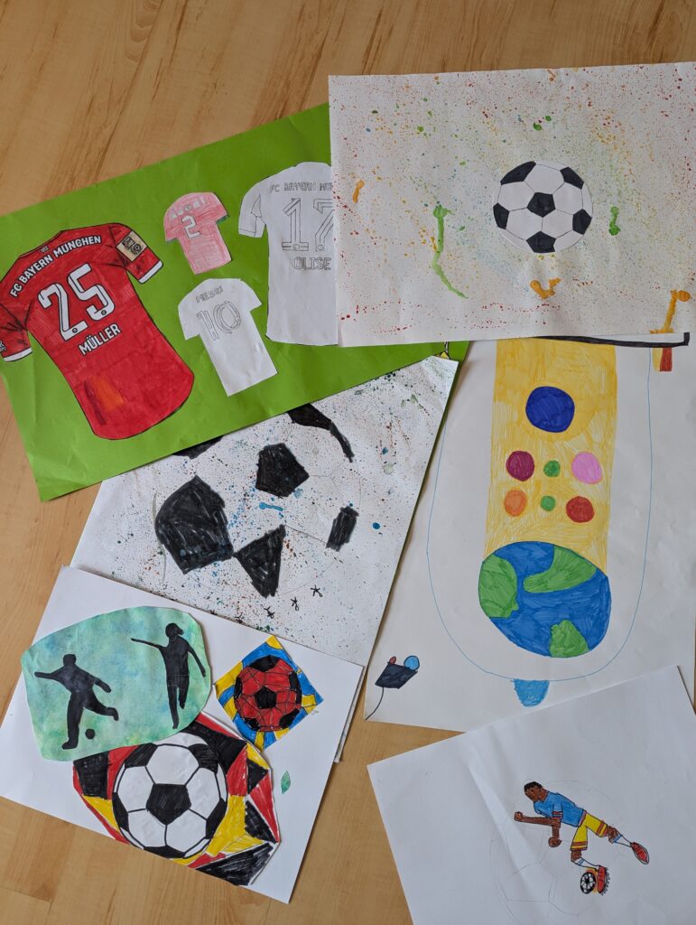 Mehrere farbenfrohe Kinderzeichnungen auf einem Holzboden mit Fußballmotiven, darunter Trikots, Fußbälle, Silhouetten von Spielern und eine große Farbpalette mit leuchtenden Farben.