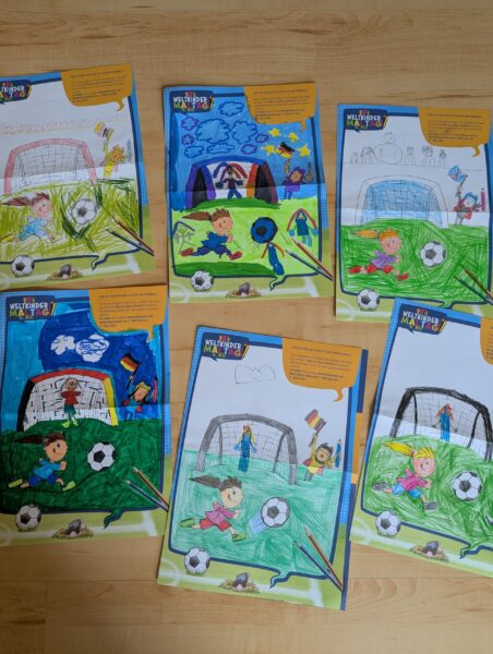 Auf einem Holzboden liegen fünf Malvorlagen, auf denen Kinder beim Fußballspielen zu sehen sind; jede ist mit Filzstiften und Buntstiften anders ausgemalt. Einige Seiten sind vollständig ausgemalt, andere hingegen nur teilweise.
