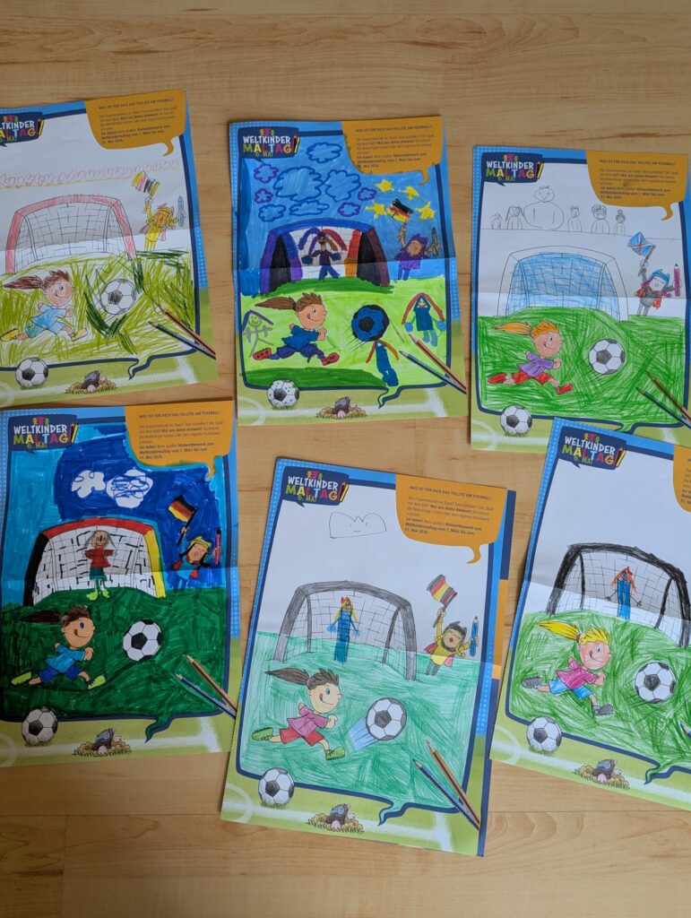 Auf einem Holzboden liegen fünf Malvorlagen, auf denen Kinder beim Fußballspielen zu sehen sind; jede ist mit Filzstiften und Buntstiften anders ausgemalt. Einige Seiten sind vollständig ausgemalt, andere hingegen nur teilweise.