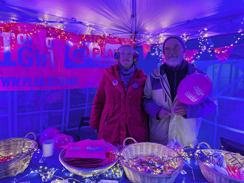 Zwei Personen stehen hinter einem Stand, der mit Lichtern und einem Plan-International-Banner geschmückt ist; der Mann hält einen rosa Luftballon in der Hand.
