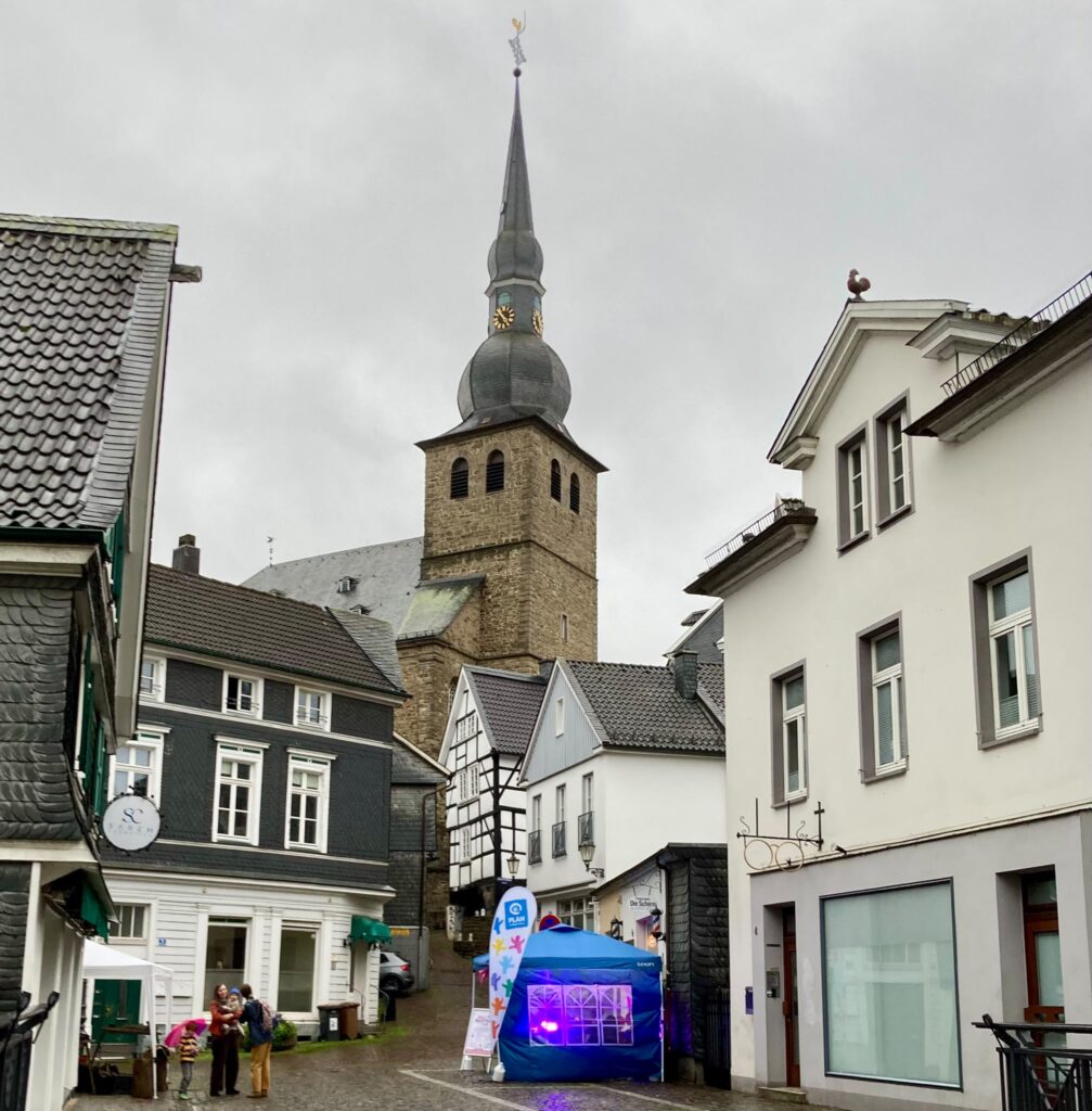 Eine schmale Straße in einer europäischen Stadt mit einem steinernen Kirchturm im Hintergrund, einem kleinen blauen Pop-up-Zelt und einigen Passanten.